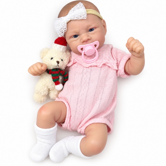 Reborn baby doll lying in pink knit romper with lace bow headband, holding Christmas teddy bear