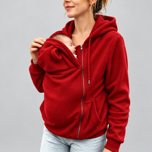 Woman wearing the red fleece kangaroo baby carrier hoodie, zip-up front pouch open with a newborn baby nestled inside, studio background